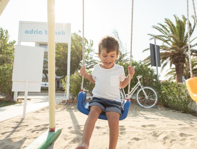 Bambino felice su un'altalena in spiaggia, vicino a una bicicletta.